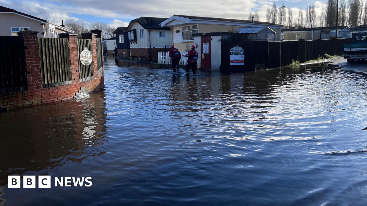 A man wearing wellies stands with his hands on his hips looking across a flooded coffee shop. The water is shallow but it is brown and covering the entire floor.