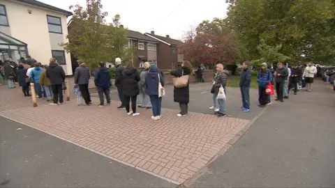 A long line of people waits single file on the side of a path, beside a patch of grass. They're waiting to get into a dentist's practice.