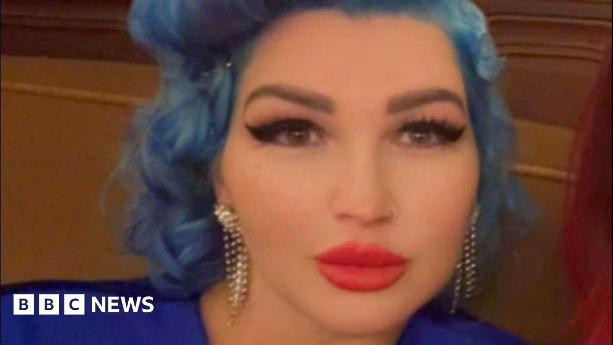 Tania Williams is inside looking to the camera. She has wavy blue hair and make-up on, including bright red lipstick and black mascara. She is wearing a dark blue dress.