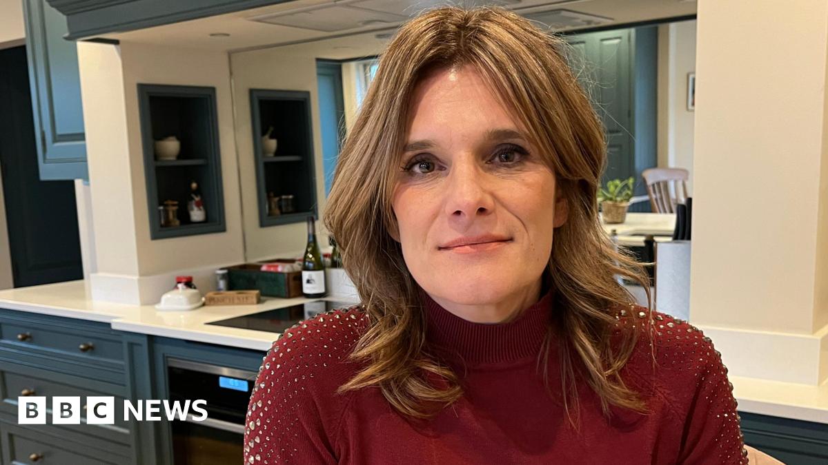 Ellen looks at the camera with a small smile. She is in a kitchen in a house, with the cabinets and work surface visible behind her. She has short dark blonde hair, which is loosely curled, and wears a red jumper with diamante sequins on the shoulders.