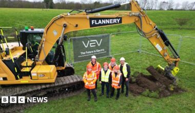 An aerial shot of six people wearing hi-vis jackets in a field, with a big yellow digger behind them.
