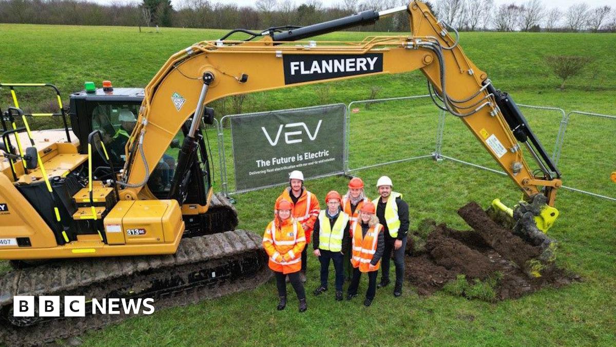 An aerial shot of six people wearing hi-vis jackets in a field, with a big yellow digger behind them.