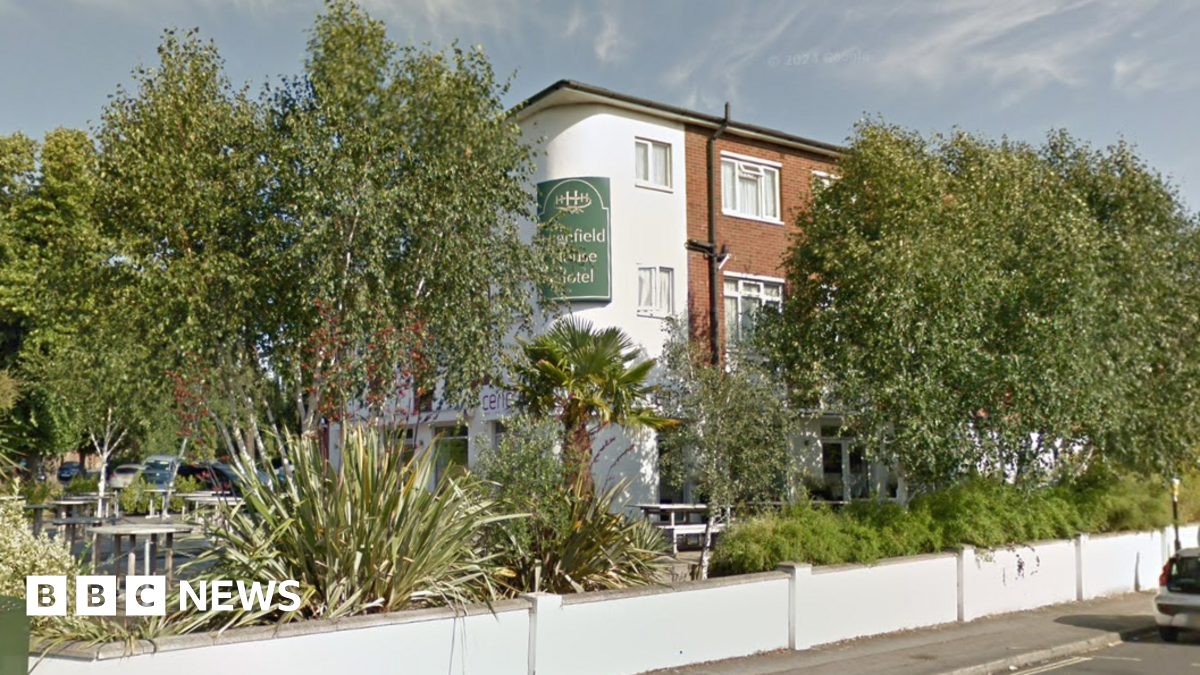 Street View image of Highfield House Hotel, a three storey building of brick and white render set back from the street corner behind a low white wall. The garden is filled with trees, palms and tropical-looking plants.
