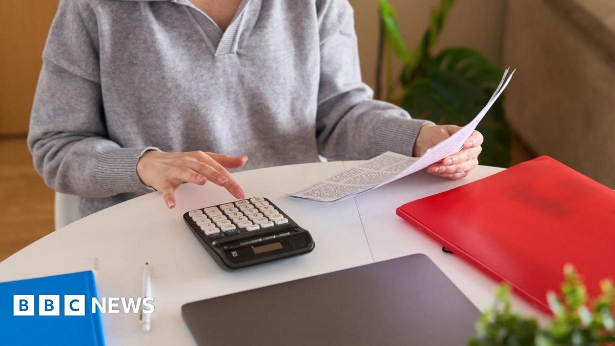 Shona Robison, who has blonde hair tied back, speaks in the Scottish Parliament chamber with her right hand held out in front of her, holding a pen. She is wearing a pink suit jacket and a black and white scarf.