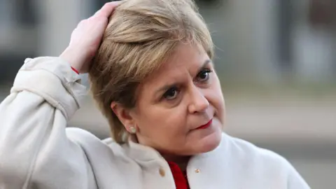 Getty Images Nicola Sturgeon standing outside, wearing a white coat and a red top. Her right arm is raised and she is touching her hair, which is short and sandy blonde. Her expression is neutral.