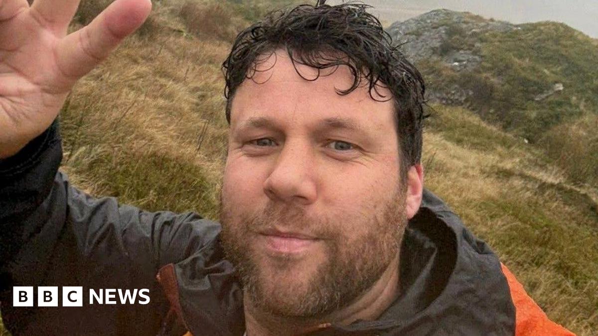 Selfie style picture of a bearded man with dark wavy hair on a hill. He is wearing a black and orange jacket and waving at the camera
