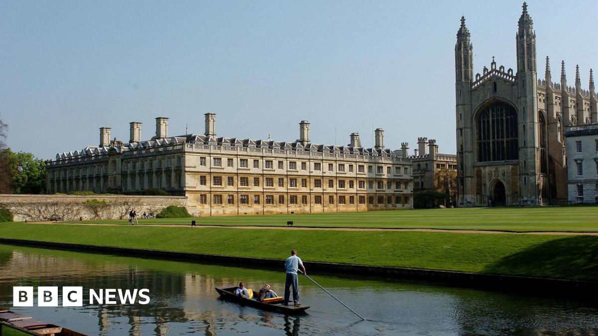 A river is in front of a large stretch of green grass that has large buildings which make up the University of Cambridge. A person is punting on the river.