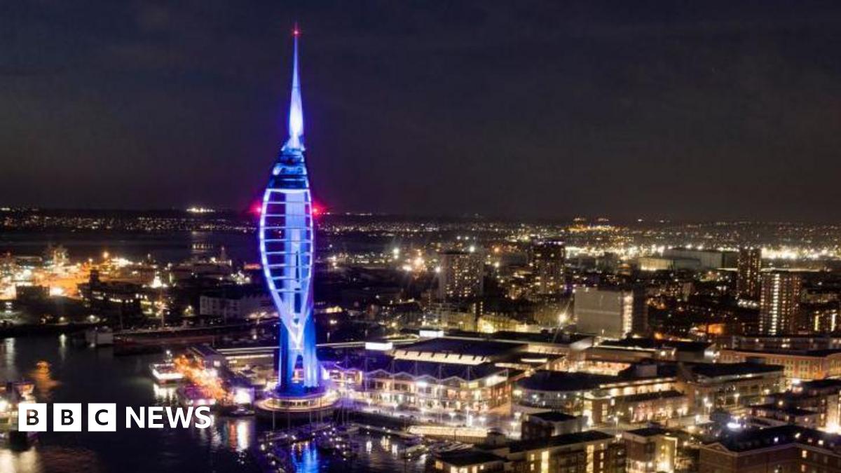 View across Portsmouth city at night. Buildings are lit up with lights whilst the sky is dark. In the foreground is the Spinnaker Tower, an iconic tall structure in the shape of a ship's sale which is illuminated blue.