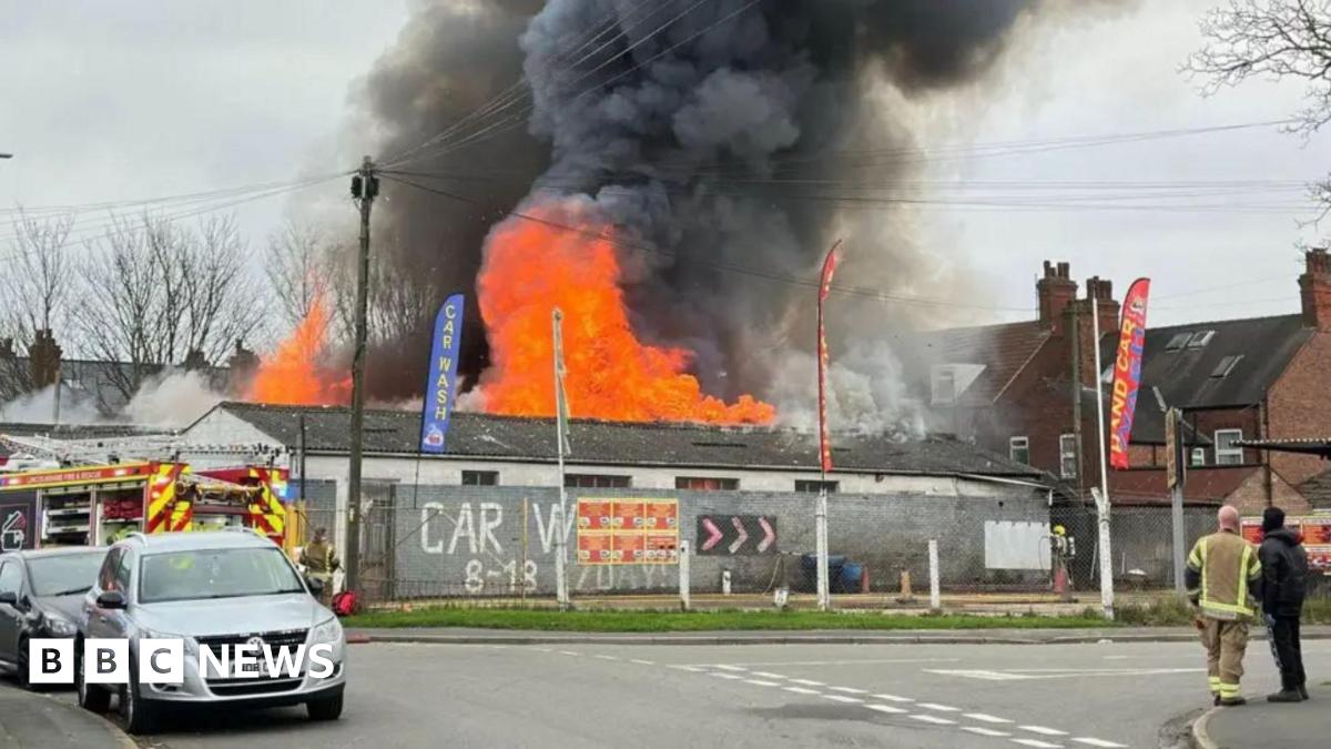 Large flames and plumes of black smoke can be seen coming from a car wash facility behind a metal fence. Emergency service workers and a fire engine are on the scene.