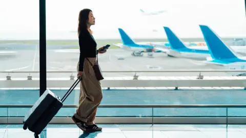 Getty Images A young woman walks in an airport with her carry-on luggage. She is carrying her phone and wearing baggy beige trousers and a black top