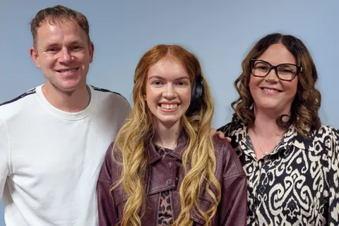 Three people standing close together against a plain light blue background. The person on the left is wearing a white long-sleeve top with dark trim on the shoulders. The person in the middle has long wavy hair and is wearing a maroon leather jacket over a patterned top. The person on the right has shoulder-length curly hair and is wearing a black-and-white animal print blouse