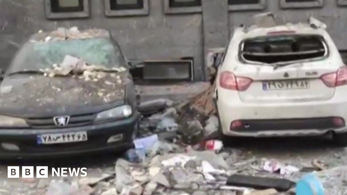 Two cars, a dark coloured Peugeot and a white hatchback both sit on the road in front of the building where the blast happened, covered in rubble