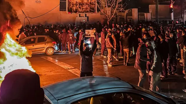 A large crowd of people wearing hoods and masks gather on the streets at nighttime in Iran. They're blocking the streets and some are holding up signs. There's a large fire to the left of the photo.