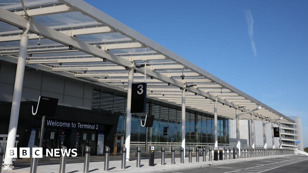 The forecourt of Manchester Airport's Terminal 2, with bollards seen running across a pedestrian area in front of parking bays. A terminal sign can be seen in front of the entrance way on a sunny day.