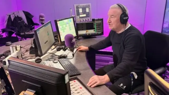 Mike has short grey hair and is wearing a black jumper. He is wearing headphones and talking into a purple microphone.