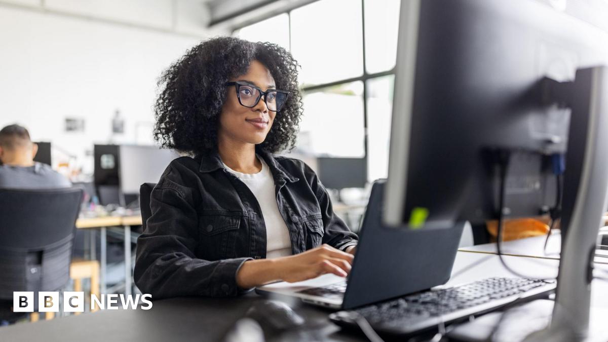 Businesswoman wearing black glasses with dark hair, sitting working on computer at her desk in office
