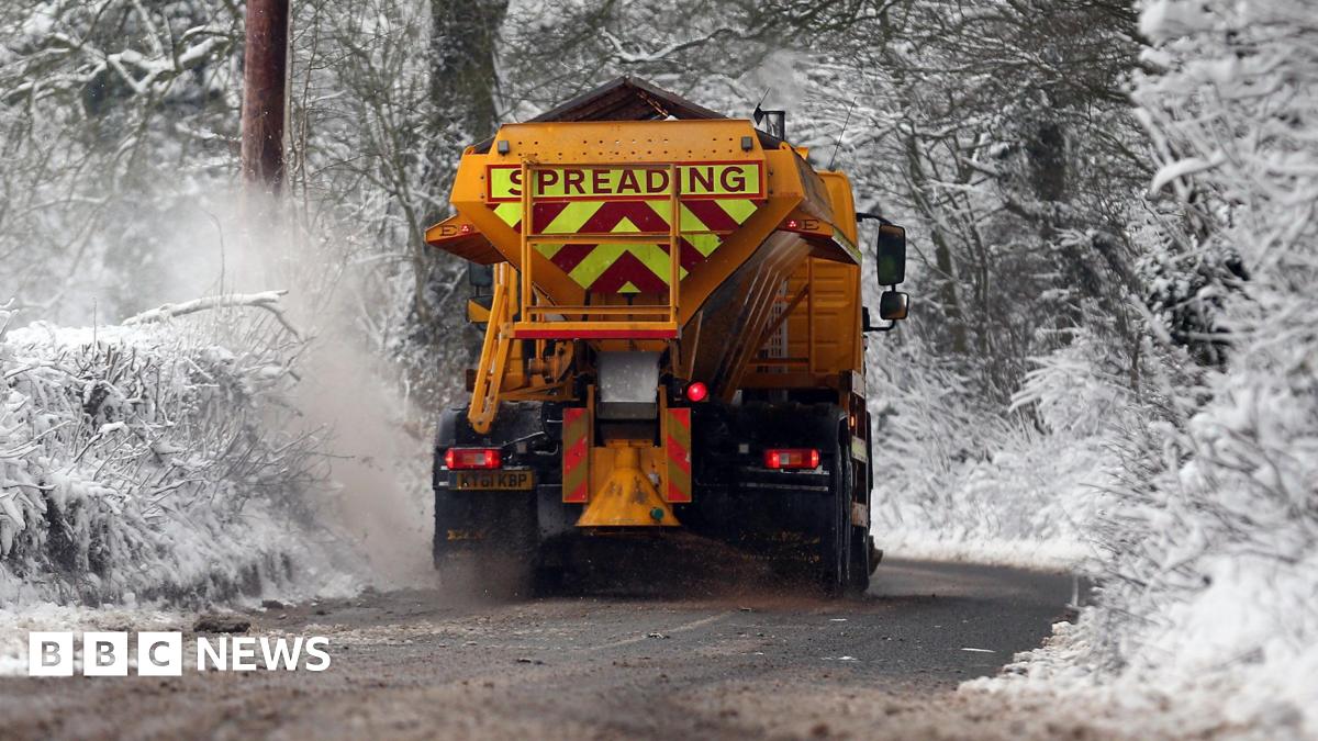 Orange gritting machine driving along snowy road, with snowy trees in the background.