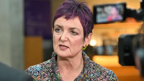 Getty Images Angela Constance, who has short purple hair, speaks to a journalist in the Scottish Parliament. She is wearing a multi-coloured jacket, with the picture taken over the shoulder of the journalist and from behind a camera.