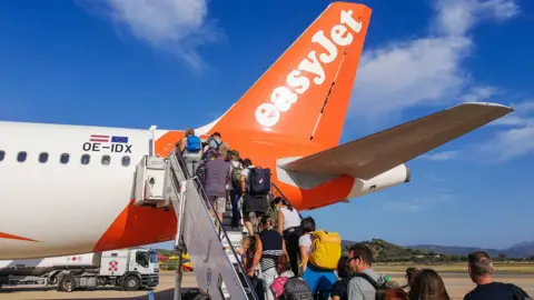Getty Images People boarding an EasyJet flight some carrying hand luggage and rucksacks on airport tarmac in Sardinia. It's a sunny day and the sky is blue. 