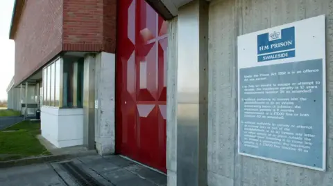 PA Media A close up image of a blue and white sign reading HM PRISON SWALESIDE, with smaller text below. It is stuck to a grey concrete wall. In the distance is a large red metal door.