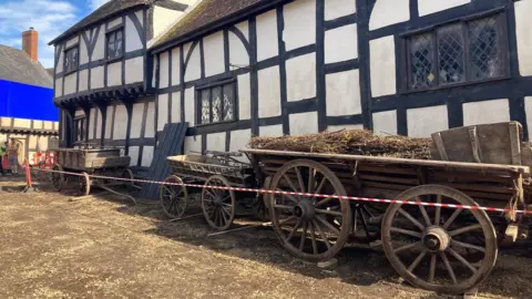 Jane Keating A black and white building with latticed windows is part of a street film set. there are old fashioned brown wood wagons next to it, and straw all over the ground