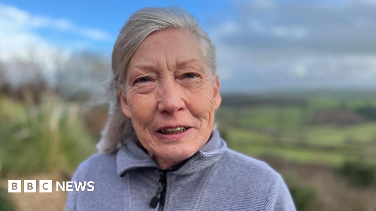 Millie Bobby Brown and David Harbour in character as Eleven and Jim Hopper. Eleven wears red shorts over grey leggings and a grey jumper. Jim Hopper wears a black leather jacket, a black beanie hat and dark trousers.They are speaking to director Frank Darabont on the set of Stranger Things. There is a vintage advertisement posted in the background with debrief on the ground.