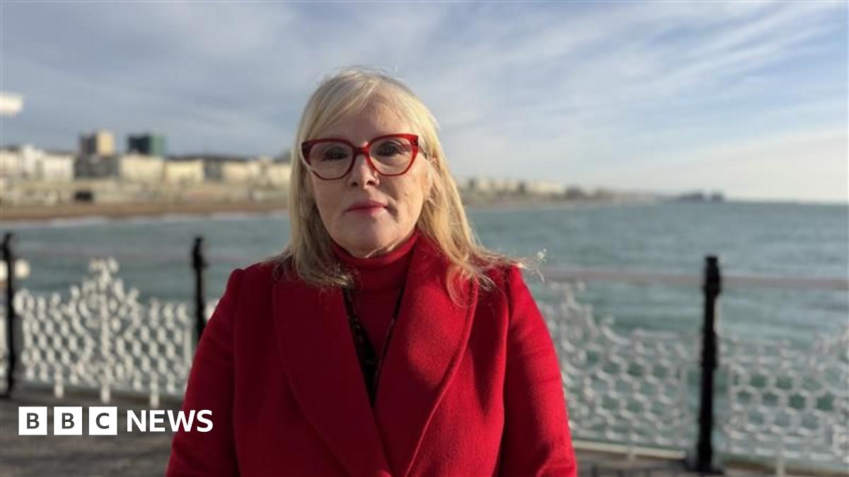 A woman wearing a red coat and red glasses. She is stood on a pier with the sea and seafront in the background.
