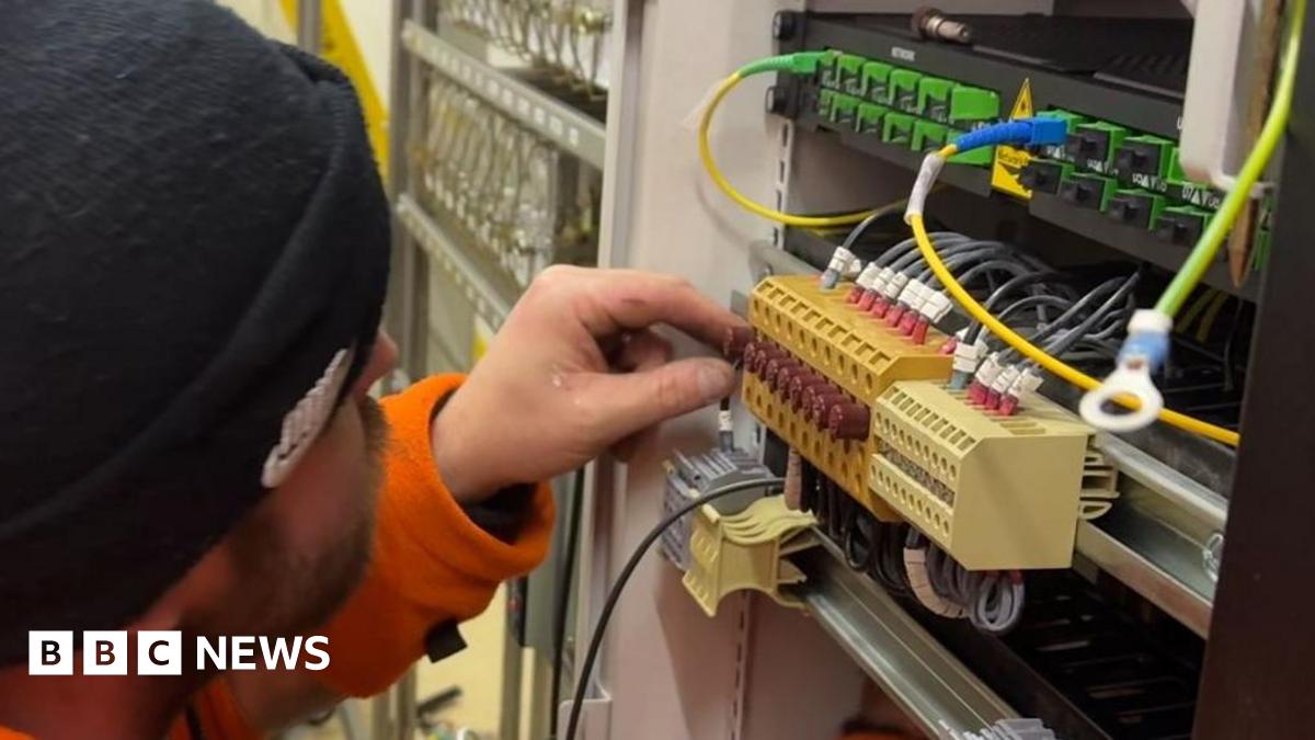 A worker wearing a black beanie and an orange high-vis uniform is working on the wires in a signalling box.