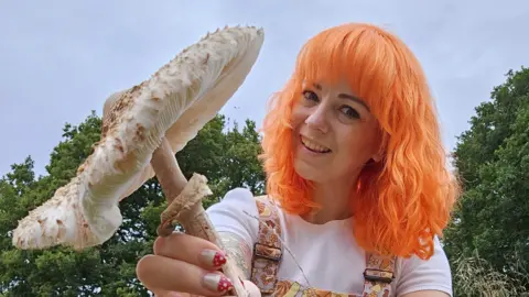Louise Gather Mrs Gather with bright orange hair holding a large white mushroom