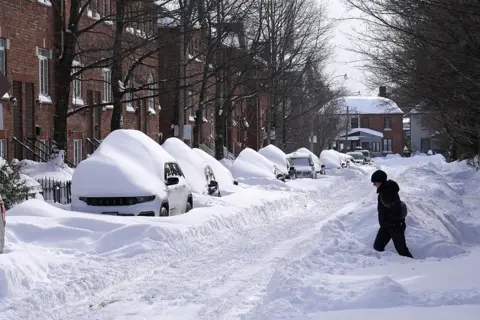 Anadolu via Getty Images A resident walks through deep snow piles in Toronto, Canada. The street is lined with cars buried under snow.