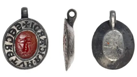 Colchester and Ipswich Museum Service Three views of an oval silver seal matrix, with a hanging loop on its top side. The first view shows the front with a thick silver rim, carved with letters and a cross at the top under the loop, while in the middle is an oval carved gemstone intaglio, showing a charioteer and two horses. The central view is of its side view, showing its thick silver rim, narrowing to a flat base. The left view is over the back, with the hanging loop at the top, with its sides narrowing to a flat base. 