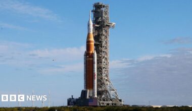 The European Service Module, which is a large cylindrical part of a spacecraft, sits in a cleanroom. On its top surface, several spherical metal tanks are surrounded by metallic insulation. There is shiny silver metallic insulation on its sides too. The module sits on a square platform, and the floor around it is marked with yellow and black safety tape.