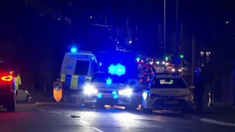 BBC Emergency service vehicles including a police van and cars on Bolton Road in Ashton-in -Makerfield in front of a line of traffic. It is dark.