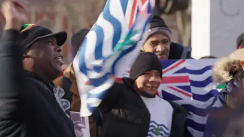 BBC A group of people protest in central London. Some are holding the flag of the British Indian Ocean Territory. 