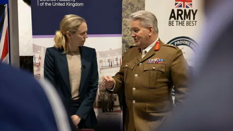 Ministry of Defence A smartly dressed women in a blue blazer jacket listens attentively as an older man in brown service dress, emblazoned with various insignia above the left breast pocket to denote his rank, speaks to her in front of a UK Army banner. 