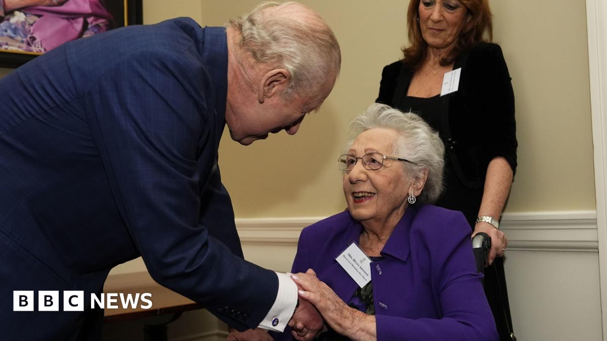 King Charles greets Helen Aronson. He wears a navy jacket and bends to shake her hand. Aronson wears a purple jacket, has short grey hair, and smiles at the King.