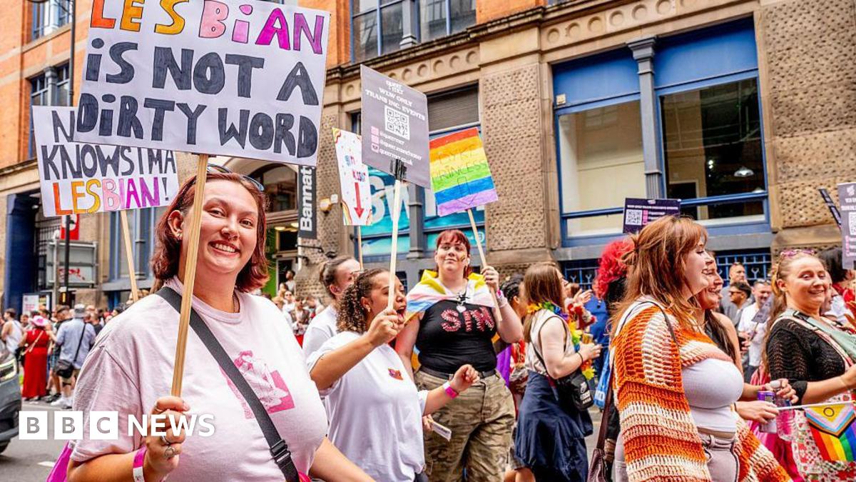 A Pride parade scene in Manchester shows people marching along the street holding signs and rainbow flags. One prominent sign reads “Lesbian is not a dirty word”. Colourful outfits and banners fill the lively crowd against a backdrop of brick buildings.