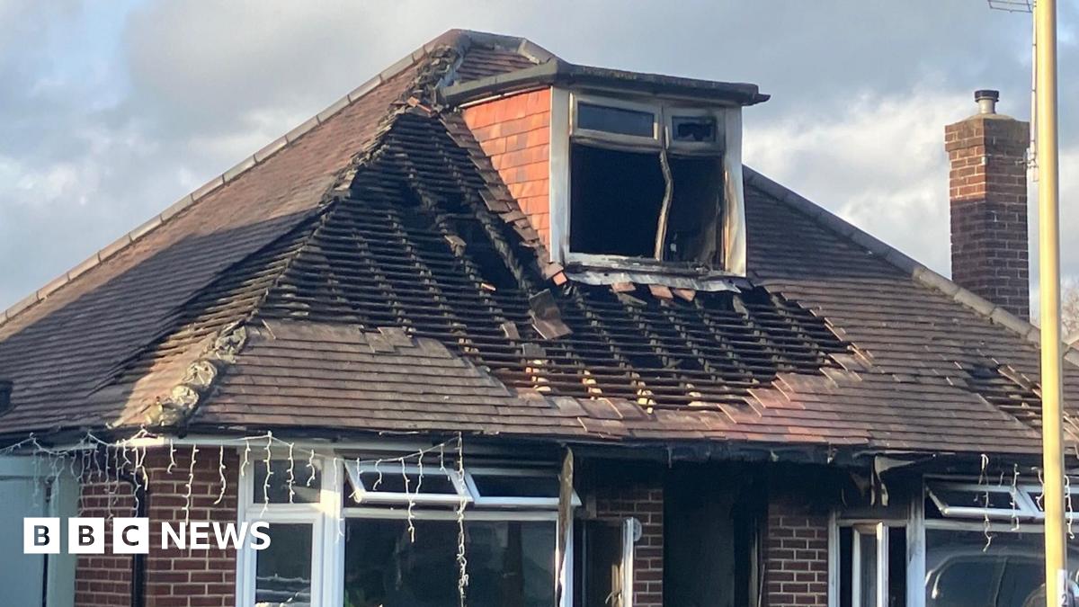 A shot of a traditional brick house with a terraced roof which has been significantly damaged by fire. Many of the roof's tiles are completely missing and a box window in the attic has been blackened and warped.
