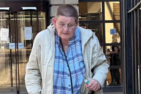 Alexander Lawrie A woman with short cropped hair and glasses, walking with the aid of a stick outside a court