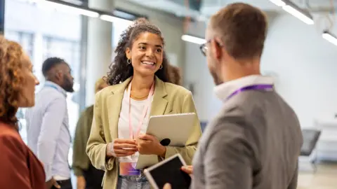Getty Images A woman with brown hair wearing a green blazer chats to a grey-haired man with grey glasses. A woman with ginger hair and a red blazer stands beside them