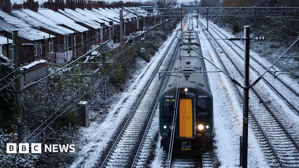 Library photograph of a black and yellow train as it travels on tracks covered in snow. Houses on the left of the tracks have snow on their roofs.