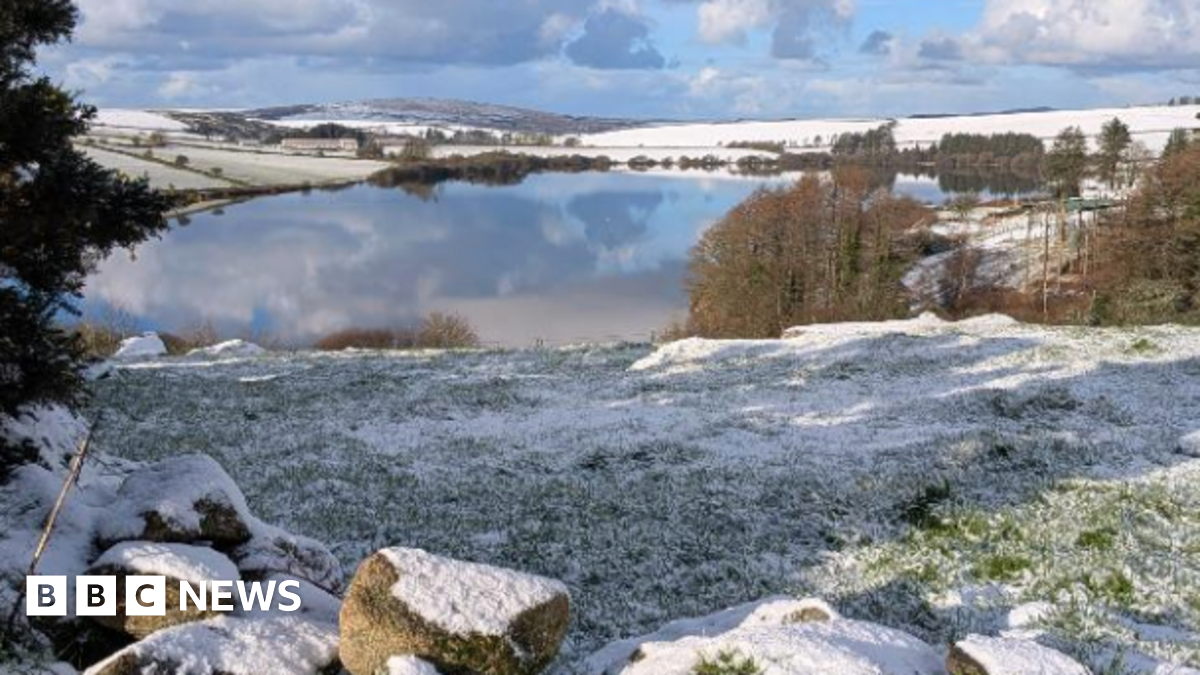 The picture shows a peaceful winter landscape with a large lake in the middle reflecting the blue sky and scattered clouds above. Surrounding the lake are gently rolling hills covered in a light layer of snow, giving the scene a crisp and bright appearance. In the foreground, there is a grassy area partially dusted with snow and a few large rocks lying near the edge. On the right side, there are clusters of leafless trees and some evergreen trees. The background stretches into distant hills, also blanketed in snow, under a sky that mixes patches of blue with dramatic white and grey clouds.
