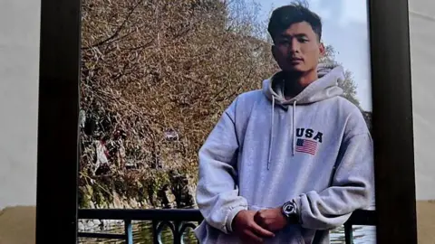 Getty Images A framed photograph of 24-year-old Anjel Chakma, wearing a hoodie and a watch, leaning against a railing, behind which there's a tree and a water body