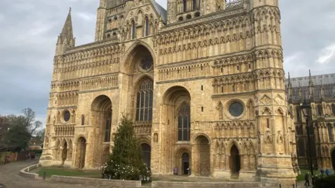 The grand, imposing west front of a medieval cathedral taken during the daytime under a cloudy grey sky. The massive, layered facade blends Norman, Romanesque and Early English Gothic styles. It has a large central arch, flanked by two smaller arches and intricate arcading. A large green Christmas tree stands In front of the cathedral.