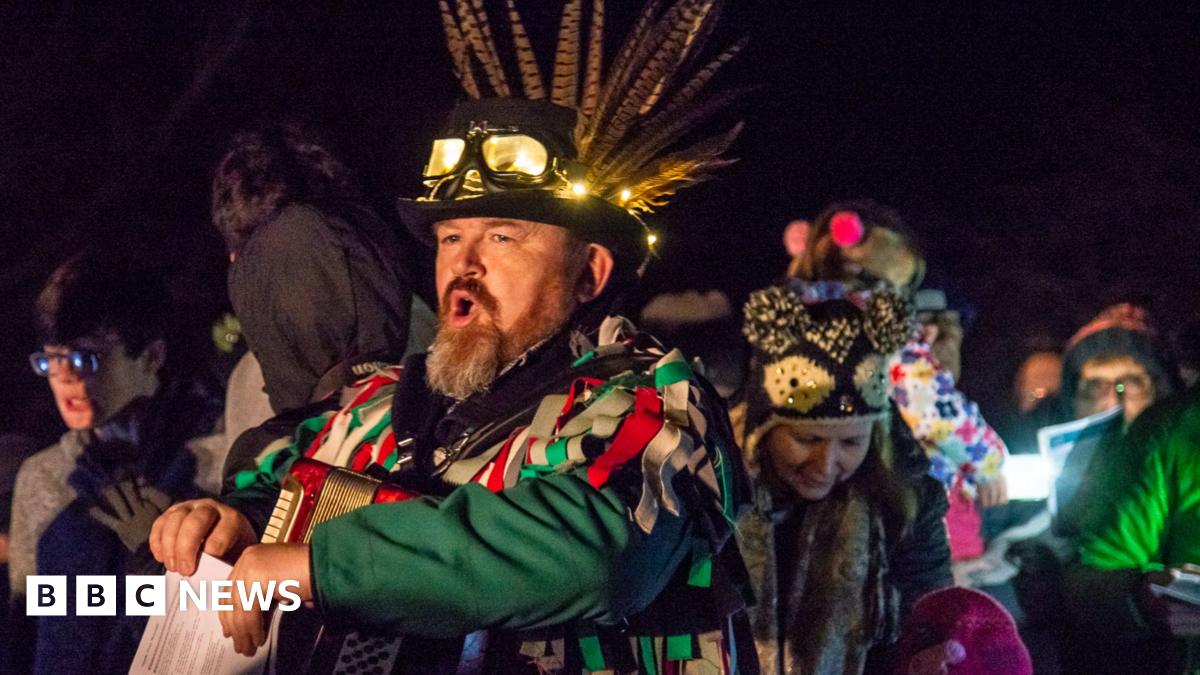 Simon processes from right to left with his mouth open as though proclaiming or singing. He wears an accordion on his chest and a feathered hat on his head. He has a green and red rag costume and holds papers that look like song lyrics. It is night time. People wearing winter coats and hats crowd around him.