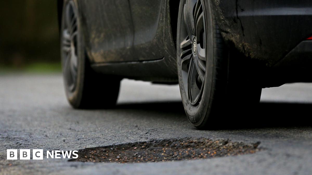 A black car parked with its rear wheel next to a large pothole in the road surface.