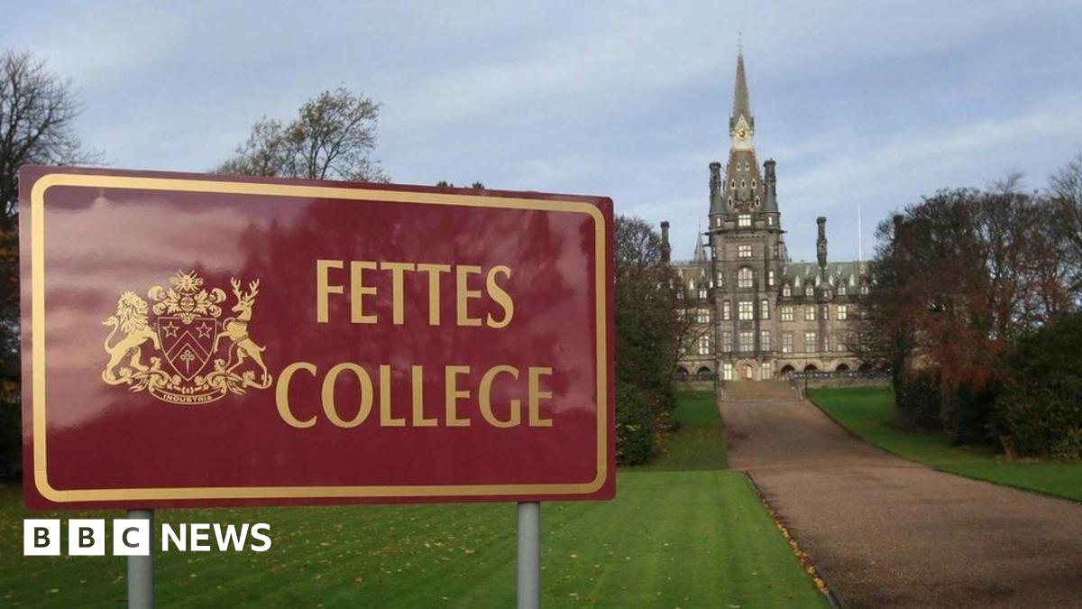 A red sign with "Fettes College" written in gold next to a coat of arms. The central turret of the stone school is visible at the end of a long pathway.