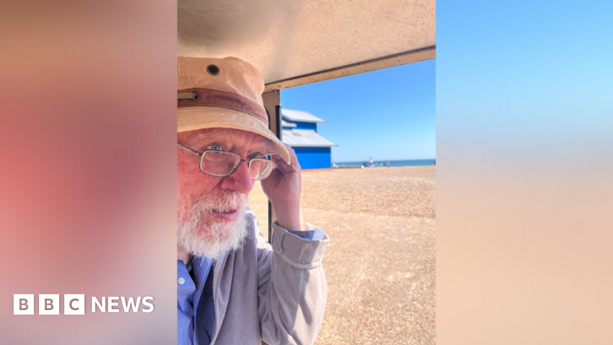 James "Max" Farrar during a trip by the sea. He is wearing a fisherman hat and glasses. The sand and see can be seen in the distance, while James is under a roof of a building or vehicle.