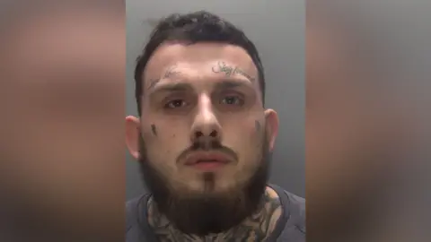 Northamptonshire Police A man with tattoos above his eyebrows and neck looks at the camera in his police custody photo. He has a thick beard.
