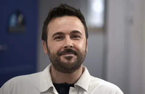 David Wilson-Wynne, man with black hair and a beard smiles at the camera. He is wearing a white shirt and black T-shirt.